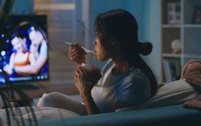 A person sitting on a couch eating noodles a television is visible in the background